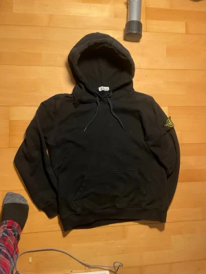 Svart hoodie från Stone Island - Svart hoodie från Stone Island med klassisk huva och dragsko. Har en stor magficka och den ikoniska Stone Island-loggan på vänster ärm. Tillverkad i mjuk bomull, perfekt för en avslappnad streetwear-look. Osäker om äkta men vid minsta fundering så är de bara o skriva 