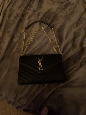  YSL väska - Snygg svart väska från YSL med klassiskt guldigt logotyp-spänne framtill. Väskan har quiltat mönster, guldfärgad kedjerem och detaljer i läder. Perfekt för att lyfta din outfit med en lyxig touch. Vet dock inte om den är äkta, har inget kvitto kvar bara kartongen, den köptes för några veckor sen så den e som ny. Priset kan diskuteras. 
