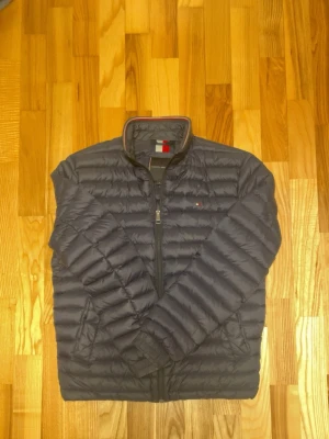 Mörkblå dunjacka Tommy Hilfiger - Snygg mörkblå dunjacka från Tommy Hilfiger med diskret logga på bröstet. Jackan har dragkedja framtill, ståkrage och två sidofickor. Lätt och smidig modell som håller värmen under kalla dagar. Klassisk design med quiltade sömmar.