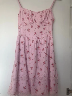 Rosa blommig klänning med spetsdetaljer - Söt rosa klänning från hollister med blommigt mönster och tunna axelband. Klänningen har volangkant och spetsdetaljer längst ner, samt smock i ryggen för skön passform. 