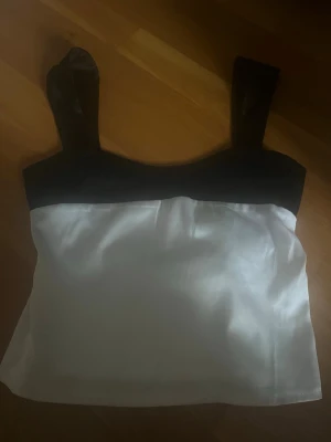 Svartvit satinlinnetopp med knytband - Snygg svartvit linnetopp i satin med breda axelband och dekorativt knytband i ryggen. Toppen passar perfekt till både jeans och kjol. Materialet ger en lyxig känsla och lätt glans.