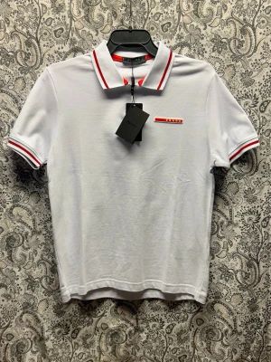 Vit Prada piké  - Vit t-shirt från Prada med klassisk pikékrage och röda ränder på krage och ärmslut. Liten röd Prada-logga på bröstet och half zip-dragkedja framtill. Tillverkad i bomull för en skön och stilren look.