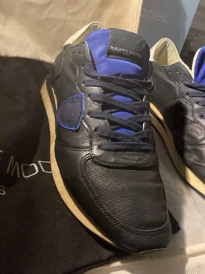 Philippe Model svarta sneakers blå detaljer - Har mejl kvitto, box och bags.Snygga svarta sneakers från Philippe Model med blå detaljer på plös och sidor. Skorna har en klassisk låg siluett, svarta skosnören och beige sula. Materialmix av läder och textil ger en exklusiv känsla och cool look.