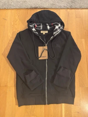Svart hoodie från Burberry - Säljer en svart hoodie från Burberry med dragkedja och klassiskt rutigt foder i huvan. Hoodien har ribbade muddar, fickor framtill och snygga detaljer. Perfekt för dig som vill ha en stilren och exklusiv look.