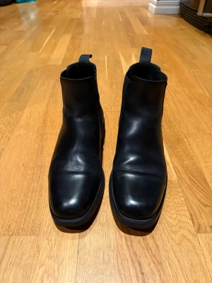 Svarta Gant Chelsea boots i skinn - Säljer ett par klassiska svarta Chelsea boots från Gant i slätt skinn med elastiska paneler på sidorna och dragflikar både fram och bak. Snygg, stilren siluett som passar till det mesta. Perfekta för dig som vill ha en tidlös och enkel look.