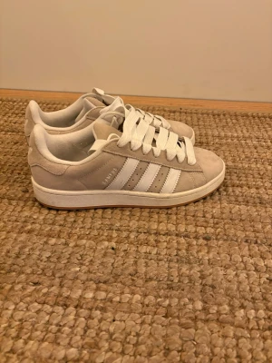 Beige Adidas Campus sneakers - Säljer ett par beige Adidas Campus sneakers i mocka med klassiska vita ränder på sidorna och vita skosnören. Skorna har en låg profil och vit sula, perfekt för en clean och stilren look. Adidas-loggan syns på sidan och hälen.