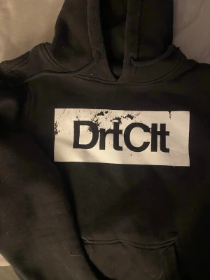 Svart DrtClt hoodie med tryck - Svart hoodie från DrtClt med stor vit logga och tryck på bröstet. Klassisk känguruficka framtill och mjuk insida. Huva med dragsko för extra komfort. Perfekt för dig som gillar streetwear och vill ha en stilren men edgy look.