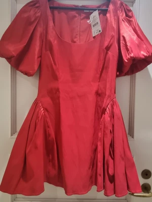 Röd kort klänning från NA-KD - Trendig röd kort klänning från NA-KD med puffärmar och vid kjol. Klänningen har en rundad urringning och är tillverkad i ett glansigt material som ger en lyxig känsla. Perfekt för dig som vill sticka ut med en färgstark och modern look.