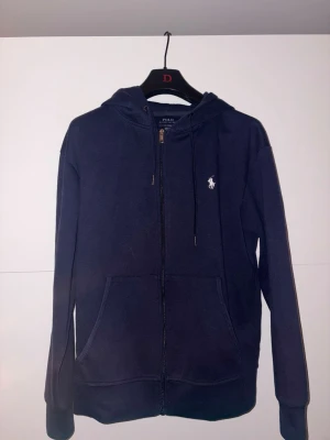 Mörkblå hoodie från Polo Ralph Lauren - Snygg mörkblå hoodie från Polo Ralph Lauren med dragkedja framtill och klassisk vit logga på bröstet. Hoodien har huva med dragsko och stora fickor framtill. Tillverkad i mjukt bomullsmaterial och har ribbade muddar vid ärmslut och nederkant. Skicka 9/10