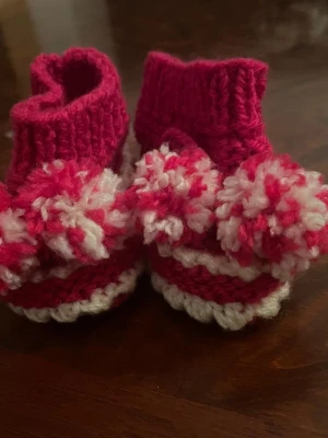 Stickade tofflor med pompoms rosa/vit - Supersöta handstickade tofflor i rosa och vitt med stora fluffiga pompoms framtill. Tofflorna har ribbstickad kant och är gjorda i mjukt garn, perfekta för att hålla fötterna varma och mysiga. Passar till dig som gillar gulliga accessoarer.