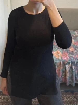 Svart ribbad långärmad topp - En svart ribbad topp med lång ärm och rund halsringning. Modellen är något längre och har en slim passform som sitter snyggt mot kroppen. Perfekt att styla med jeans eller kjol för en clean look.