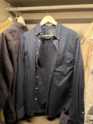 Mörkblå skjorta från Jack and Jones  - Klassisk mörkblå skjorta från Jack and Jones med knappstängning och bröstficka. Skjortan har lång ärm och button-down krage. Tillverkad i mjuk bomull som känns skön mot huden. Perfekt för en avslappnad men ändå stilren look. Använd en gång.