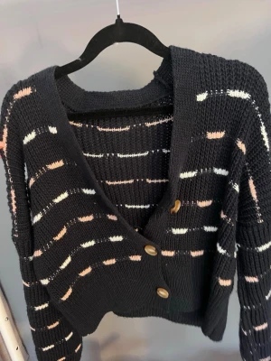 Svart stickad kofta med knappar - Snygg svart stickad kofta med v-ringning och tre träknappar framtill. Koftan har ett mönster med vita och ljusrosa ränder och är lite croppad i modellen. Perfekt att slänga över en topp för en avslappnad look. Köpt i Spanien 