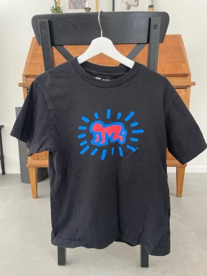 Svart Uniqlo UT Archive t-shirt - Säljer en svart t-shirt från Uniqlo UT Archive med ett färgstarkt tryck framtill i blått och rött. T-shirten har rund hals, korta ärmar och är gjord i mjuk bomull. Perfekt för dig som gillar streetwear och grafiska prints.