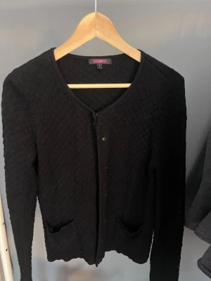 Svart quiltad kofta från Indiska - Snygg svart kofta från Indiska med quiltat mönster, rund halsringning och knäppning framtill. Koftan har två små fickor fram och långa ärmar. Perfekt för dig som gillar stilrena och bekväma plagg.