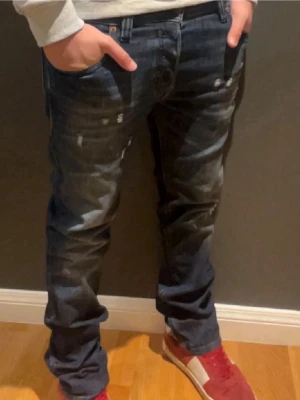 Mörkblå slitna jeans med tvättade detaljer - Säljer ett par mörkblå jeans med tvättade och slitna detaljer samt synliga sömmar. Jeansen har fem fickor, raka ben och en avslappnad passform. Materialet är klassisk denim i bomull. Perfekta för en casual och trendig look.