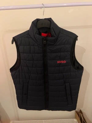 Väst från HUGO - Snygg marinblå dunväst från HUGO med röd logga på bröstet. Västen har hög krage, dragkedja framtill och två fickor med knappstängning. Quiltad design och lätt vadderad för extra värme. Perfekt för lager-på-lager och streetwear-stil.