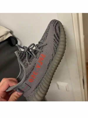 Yeezy Boost 350 V2 Beluga - Grymma Yeezy Boost 350 V2 Beluga med grå Primeknit-överdel, orange 'SPLY-350' text på sidan och zebramönstrade detaljer. Skon har en genomskinlig, ribbad sula och matchande grå skosnören. Perfekt för dig som vill sticka ut med stilrena sneakers.