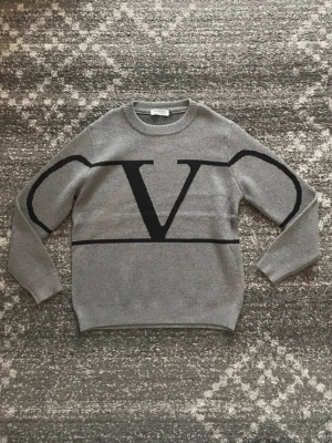 Grå stickad tröja från Valentino - Snygg grå stickad tröja från Valentino med stor svart V-logga framtill och svarta detaljer på ärmarna. Tröjan har rund halsringning, långa ärmar och ribbade muddar. Perfekt för dig som gillar stilrena och exklusiva plagg.