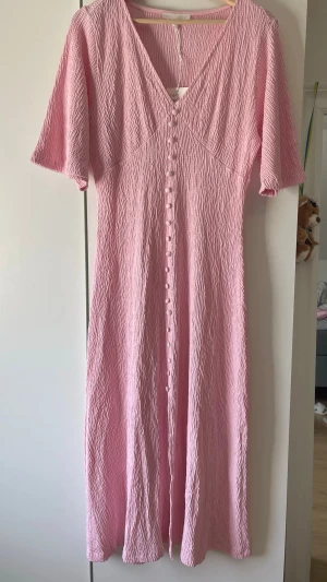 Ljusrosa långklänning med knappar - Säljer en ljusrosa långklänning med korta ärmar och knappar hela vägen framtill. Klänningen har en mjuk, flowy passform och är perfekt för varma dagar. Ingen synlig logga eller märke.