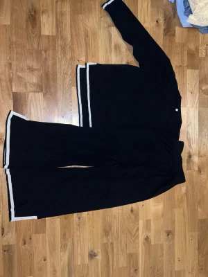 Svart loungewear-set med vita ränder - Mysigt svart loungewear-set med långärmad tröja och vida byxor. Båda plaggen har vita ränder längs kanterna för en stilren look. Perfekt för avslappnade dagar hemma.