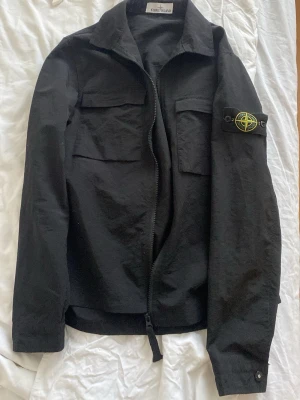 Svart overshirt från Stone Island - Snygg svart overshirt från Stone Island med klassisk patch på vänster ärm. Jackan har två bröstfickor med lock, dragkedja framtill och knappdetaljer vid ärmslut. Tillverkad i ett slitstarkt bomullsmaterial och har en rak passform som ger en clean look.