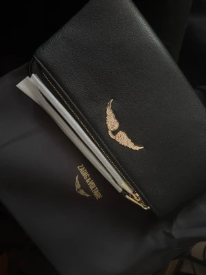 Svart kuvertväska Zadig & Voltaire - Stilren svart kuvertväska i läder från Zadig & Voltaire med guldfärgad kedja och ikonisk vingdetalj framtill. Väskan har dragkedja och en lyxig känsla med detaljer i guld. Perfekt för att lyfta din outfit med en edgy touch.