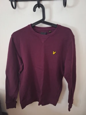 Vinröd sweatshirt från Lyle & Scott - Säljer en vinröd sweatshirt från Lyle & Scott med klassisk rund halsringning och gul logga på bröstet. Tröjan har ribbade muddar vid ärmslut och nederkant. Perfekt för dig som gillar stilrena och bekväma plagg.