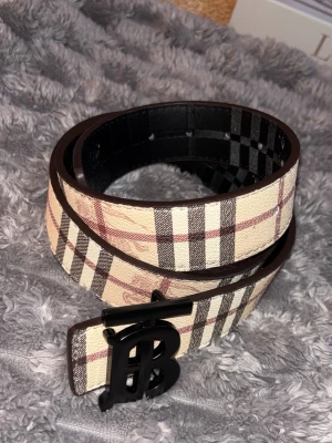 Rutmönstrat bälte från Burberry - Snyggt bälte från Burberry med klassiskt beige, svart och rött rutmönster. Bältet har ett stort svart metallspänne med Burberrys ikoniska monogram. Perfekt accessoar för att lyfta din outfit.