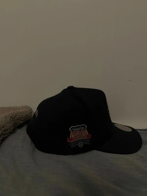 Svart keps Mitchell & Ness 120 Years - Snygg svart keps från Mitchell & Ness med broderad text bak och ett färgglatt 120 Years-märke på sidan. Lite nopprig, lite dammig men jag fixar såklart det innan jag skickar den