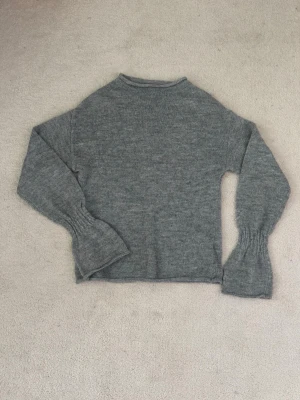 Grå stickad tröja med vida ärmar - Mysig grå stickad tröja med rund hals och långa, lätt utsvängda ärmar. Tröjan har en enkel och stilren design, perfekt för lager på lager och passar till det mesta. 