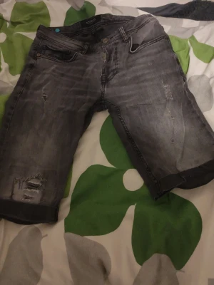 Grå jeansshorts från Icono Milano - Säljer ett par grå jeansshorts från Icono Milano med slitna detaljer och råa kanter. Shortsen har fem fickor, knappgylf och en klassisk passform. Perfekta för dig som gillar en avslappnad och trendig stil.