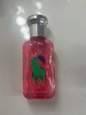 Ralph Lauren Big Pony Pink Parfym - En cool parfym från Ralph Lauren Big Pony-serien i en genomskinlig rosa glasflaska med silverfärgat lock. Flaskan har en grön polospelare på framsidan och en stor blå siffra 2 på baksidan. Perfekt för dig som gillar färgstark och sportig design.