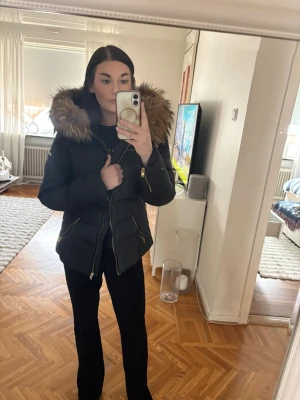 Svart dunjacka med pälskrage från Rockandblue - Säljer en svart dunjacka från Rockandblue med stor, fluffig fuskpälskrage i brunt. Jackan har guldiga dragkedjor, flera fickor och Rockandblue-logga på ärmen. Snygg, figursydd modell som håller dig varm under kalla dagar. Finns ett litet hål på armen (se bild) finns lätt att köpa svart jacktejp för att klistra på