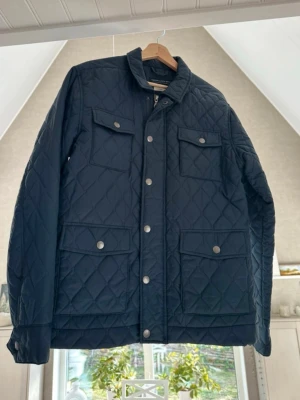 Jack&Jones quiltad jacka  - Snygg marinblå quiltad jacka med fyra framfickor och tryckknappar. Jackan har ståkrage och dragkedja samt knäppning framtill. Perfekt för kyliga dagar och ger en stilren look. Materialet är lätt och bekvämt, med klassiskt rutmönster.