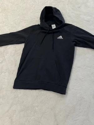 Svart hoodie från Adidas med logga - Svart hoodie från Adidas med vit logga på bröstet och tre vita ränder på ena ärmen. Klassisk känguruficka framtill och justerbar huva med snören. Tillverkad i mjukt bomullsmaterial för skön känsla. Perfekt för en avslappnad och sportig stil.