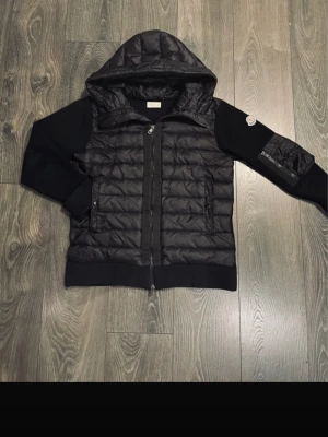 Svart cardigan från Moncler - Snygg svart hybridjacka från Moncler med quiltad front och huva i dun, samt stickade ärmar och rygg. Jackan har dragkedja framtill, fickor med dragkedja och Moncler-logga på ena ärmen. Perfekt för dig som vill ha en sportig men stilren look.