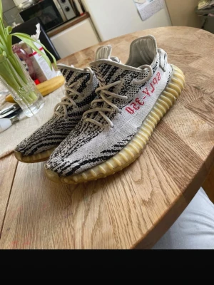 Yeezy Boost 350 V2 Zebra sneakers - Säljer ett par Adidas Yeezy Boost 350 V2 Zebra med vit och svart randigt Primeknit-tyg, vita snören och den klassiska genomskinliga sulan. På sidan står det 'SPLY-350' i rött. Perfekta för dig som gillar streetwear och vill sticka ut.