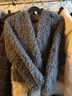 Gråbrun fluffig pälsväst - Säljer en supermjuk och fluffig pälsväst i gråbrunt med ljusare beige nederkant. Västen har en oversized krage och är gjord i fuskpäls som ger en lyxig känsla. Perfekt att slänga över en hoodie eller tröja för extra stilpoäng.