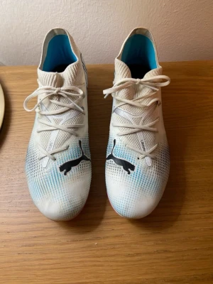 Puma vita fotbollsskor med dobbar - Snygga vita fotbollsskor från Puma med svarta loggor och blå detaljer. Skorna har snörning och en ovandel i syntetmaterial. Sulan är rosa med fasta dobbar för bra grepp på planen. Insidan är blå för extra stilpoäng.