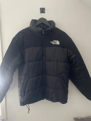 Svart pufferjacka från The North Face - Säljer en svart pufferjacka från The North Face med hög krage och dragkedja framtill. Jackan har logga på bröstet och ryggen, samt justerbar i nederkant. Perfekt för kylan