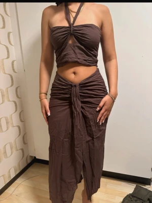 Brun bandeau topp med halterneck - Säljer en brun bandeau topp med halterneck och twistad detalj framtill och kjolen till. Toppen är ärmlös och croppad, perfekt för sommaren. Materialet känns mjukt och lätt, troligen bomull eller polyester. Snygg och trendig modell som passar till många outfits. Kjolen är omlott framtill. Säljer alltid hela setet. Toppen är i S och kjolen i M.