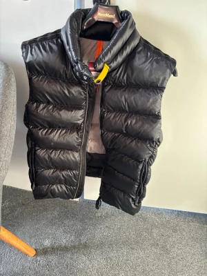 Svart dunväst från Parajumpers - Snygg svart dunväst från Parajumpers med hög krage och gul detalj vid dragkedjan. Västen har en quiltad design och logotyp på ryggen. Perfekt för lager-på-lager och håller dig varm med sin fluffiga dunfyllning.