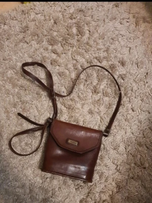 Brun axelväska i läder - Snygg brun axelväska i äkta läder med justerbar rem och klassisk fyrkantig form. Väskan har ett lock med metallspänne framtill och en praktisk dragkedjeficka på baksidan. Perfekt för dig som gillar stilrena accessoarer med vintagekänsla.