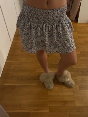 Lila blommig kjol från Gina Tricot - Kort kjol från Gina Tricot i lila, svart och ljusgul med blommigt mönster. Kjolen har bred smock i midjan och volangig nederdel som ger en söt och avslappnad look. Perfekt för dig som gillar en feminin och trendig stil.