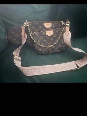 Louis Vuitton Monogram axelväska - Snygg axelväska från Louis Vuitton i klassiskt brunt monogrammönster med guldiga detaljer och kedja. Väskan har bred beige axelrem och extra liten rund väska på sidan. Perfekt för dig som gillar lyxig och trendig design.