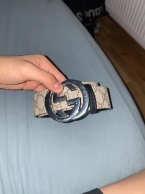 Beige GG-bälte från Gucci - Snyggt beige bälte från Gucci med klassiskt GG-mönster och stor silverfärgad GG-logga som spänne. Bältet är gjort i canvas med läderdetaljer och har en riktigt ikonisk look som lyfter vilken outfit som helst. Den är 106,5cm lång med metal saken