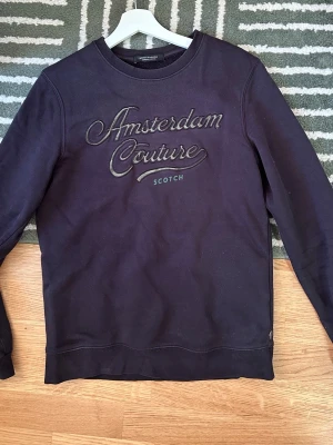 Mörkblå sweatshirt från Scotch & Soda - Mörkblå sweatshirt med broderad text 'Amsterdam Couture' och 'Scotch' på bröstet. Tröjan har rund halsringning och är tillverkad i mjukt bomullsmaterial. Perfekt för en avslappnad och stilren look.