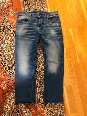 G-Star Jeans med feta fades - Säljer ett par blå jeans med snygga slitningar och tvättade detaljer på både fram- och baksida. Jeansen har fem fickor, raka ben och klassisk knappgylf. Perfekta för en avslappnad och trendig look. W33 L30