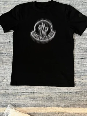 Svart Moncler t-shirt med logga - Snygg svart t-shirt från Moncler med ett stort, vitt Moncler-logotyp på bröstet samt ärmen. Klassisk rund halsringning och korta ärmar. Perfekt för dig som gillar stilrena plagg med exklusiv känsla. PRIS KAN DISKUTERAS 